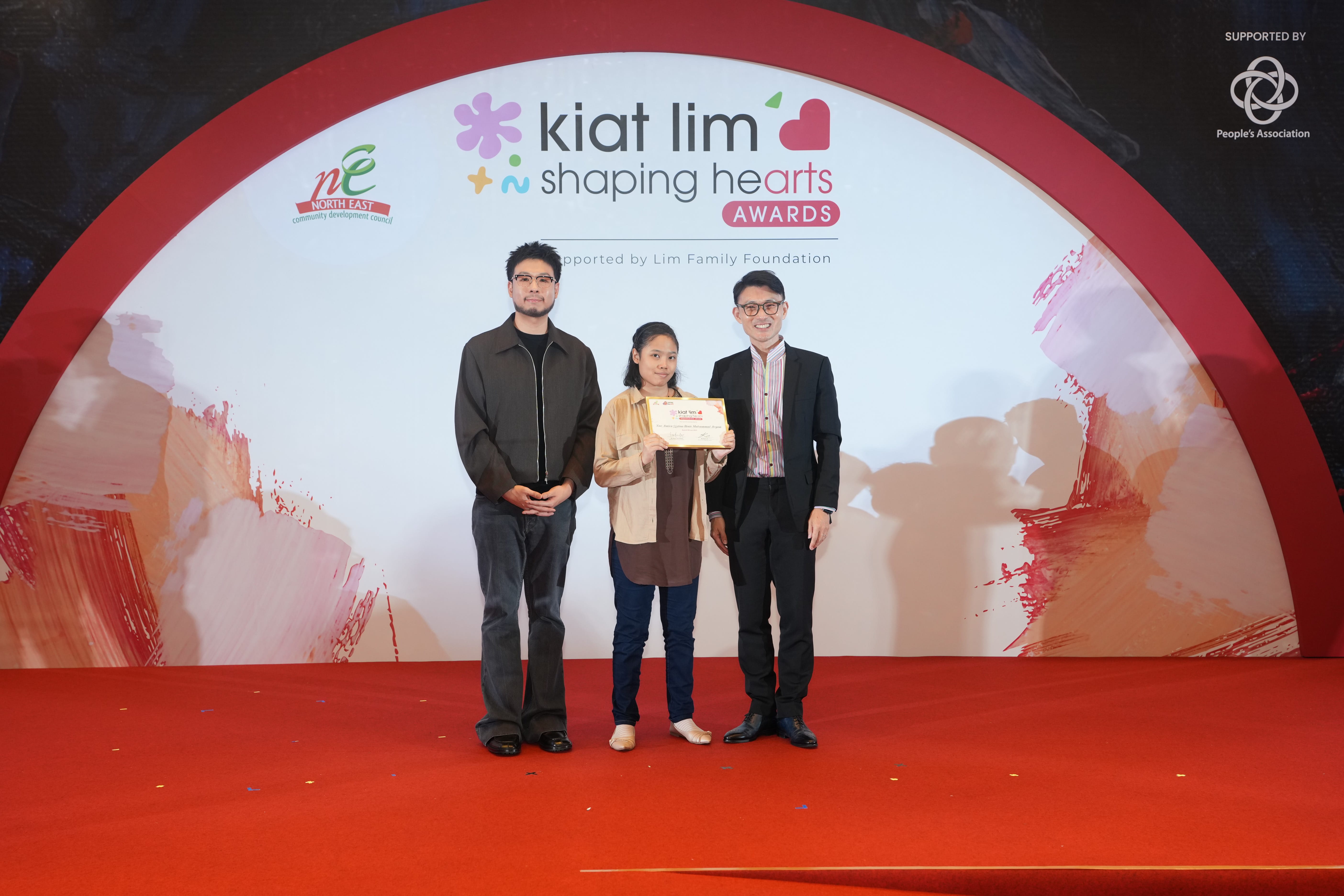 Award recipient standing on stage with the presenter, holding their certificate and posing for a group photo during the award ceremony, with a backdrop reading ‘Kiat Lim-Shaping Hearts Awards'.